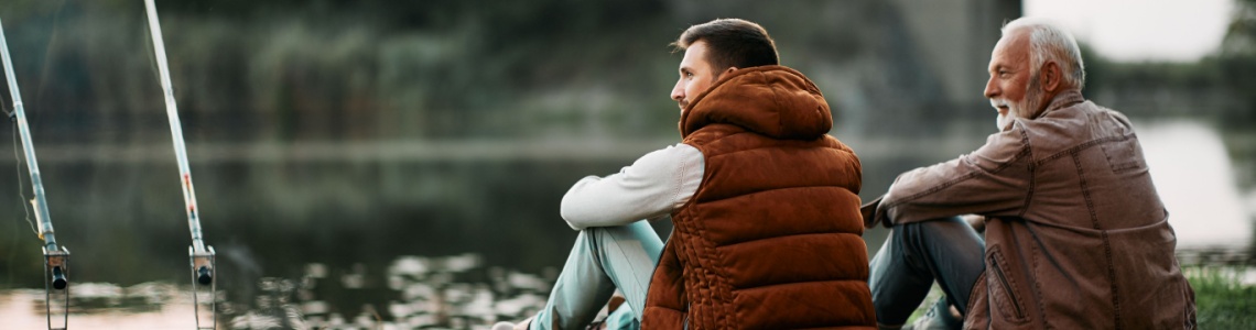 Two men sitting together and fishing by a river.